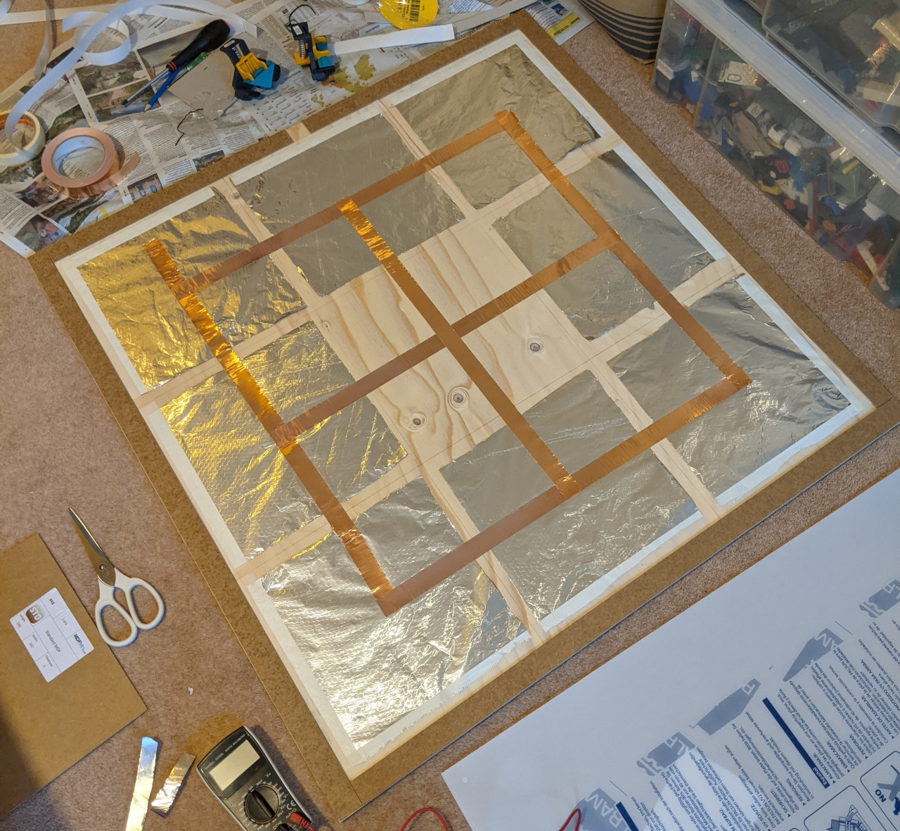 A piece of wood with some aluminium foil and copper tape on it. Tools, bits of newspaper, and backing from the copper tape are strewn around.